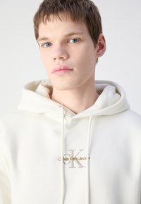 Sudadera blanca hecha de tela suave, con un bolsillo tipo canguro en el frente y un logotipo bordado en oro a tono. Presenta cordones en la capucha.