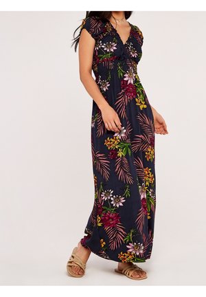 Frau trägt ein marineblaues Maxikleid mit buntem tropischem Blumenmuster und kurzen Ärmeln, kombiniert mit beigen flachen Sandalen.