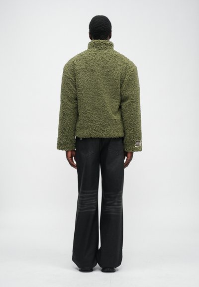 Green fleece jacket with a high collar, textured surface, and oversized fit. Paired with dark wide-leg pants.