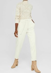 Kvinna i en krämfärgad mönstrad polotröja, högmidjade krämfärgade byxor med dragsko och beiga ankelstövlar med klack.