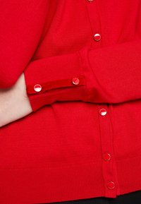 Röd stickad cardigan med texturerad tyg, runda röda knappar och en åtsittande ärm med en subtil manschettdetalj.