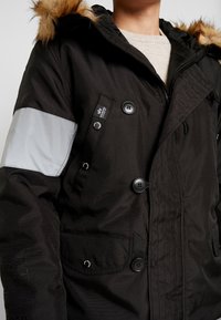 Veste d'hiver noire avec capuche bordée de fourrure, grands boutons, fermeture éclair et à boutons, et une bande grise réfléchissante sur le bras gauche.