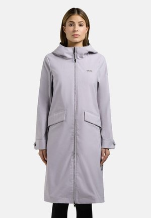 Kvinne iført lysegrå lang hettejakke med frontglidelås og klafflommer, stående foran en ensfarget hvit bakgrunn.