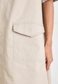 Peça de vestuário bege com um grande bolso frontal angular com fecho de aba. O tecido parece suave e leve.