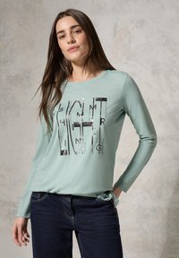 Helles grünes langärmliges T-Shirt mit Rundhalsausschnitt und einem grafischen Druck, der das Wort "LIGHT" in Schwarz und metallischen Akzenten zeigt. Kombiniert mit dunklen Jeans.