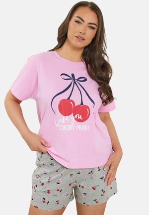 Kobieta w różowej koszulce z czerwonym nadrukiem wiśni i napisem "Kocham cię bardzo jak wiśnię!" oraz szarych szortach w wzór czerwonych i różowych wiśni.