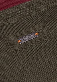 Mörkgrön stickad tyg med en liten rektangulär metalltag som är sydd med orange tråd och visar "BOSS HUGO BOSS" logotyp.