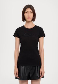 Maglietta nera in cotone, con maniche corte e collo rotondo. La parte frontale presenta un design del logo lucido e testurizzato in nero.