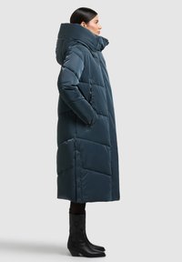 Dlouhý nadýchaný tmavě petrolejový kabát s vysokým límcem, obsahující prošívané části, boční kapsy a cvočky, doplněný černými botami.