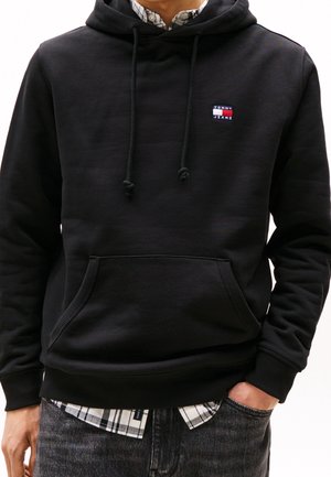 Homme portant un sweat à capuche noir Tommy Jeans par-dessus une chemise à carreaux, une main dans la poche et un jean foncé.
