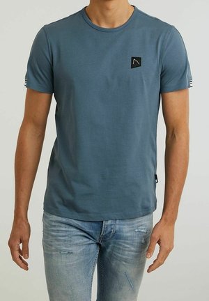 Mężczyzna w prostym niebieskim t-shircie z krótkim rękawem, z małą czarną łatką na lewej piersi oraz w jasnoniebieskich znoszonych dżinsach.