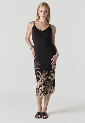 Mujer rubia con vestido midi negro sin mangas con estampado botánico beige en el dobladillo, llevando sandalias negras tipo chancla y pulseras plateadas.