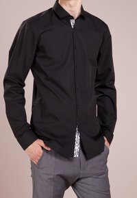 Mann trägt ein schwarzes Hemd mit Knöpfen über einem weißen, gemusterten Unterhemd, kombiniert mit grauer Hose, Hände in den Taschen, vor einfachem Hintergrund.