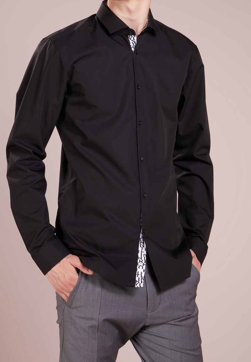 Mann trägt ein schwarzes Hemd mit Knöpfen über einem weißen, gemusterten Unterhemd, kombiniert mit grauer Hose, Hände in den Taschen, vor einfachem Hintergrund.