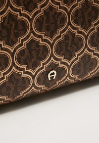 Brązowa torebka w geometryczny wzór, z złotymi elementami metalowymi i wyraźnym, wytłoczonym logo marki na powierzchni.