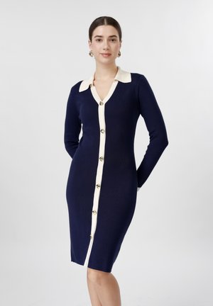 Kvinne stående iført en tettsittende marineblå kjole med hvit krage og knapperekke, hendene bak ryggen, nøytral grå bakgrunn.