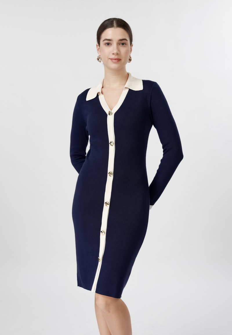 Femme debout portant une robe bleu marine ajustée avec un col blanc et une bande de boutons, mains derrière le dos, fond gris neutre.