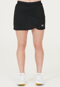 Kvinna klädd i en svart sportkjol med "FZ"-logotyp och vita sportskor, stående framför en enfärgad vit bakgrund.