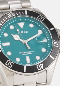 Reloj de acero inoxidable plateado con una esfera turquesa, bisel negro, marcadores de horas blancos y una ventana de fecha. Resistente al agua hasta 100m.