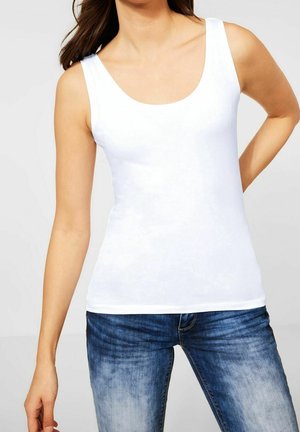 Woman wearing a plain white sleeveless tank top paired with faded blue jeans, standing with one hand on her hip.