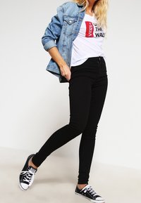 Černé úzké džíny ve spojení s bílým tričkem s potiskem a světlou denimovou bundou. Černé tenisky dotvářejí celkový vzhled.