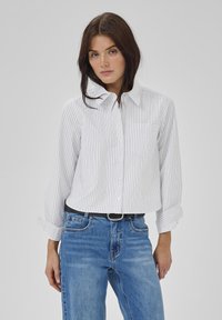 Camicia bianca a maniche lunghe con sottili righe verticali nere, dotata di colletto, taschino sul petto e maniche arrotolate, abbinata a jeans blu e una cintura nera.