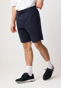 Pantalones cortos azul marino con cintura elástica y cordón, con bolsillos laterales y un corte relajado. Combinados con calcetas blancas y zapatillas negras.