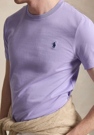 Férfi világoslila, testhezálló, rövid ujjú pólóban, melyen sötét hímzett polo játékos logó található a mellkasán, bézs színű kötött pulóverrel, amelyet derék köré kötött.