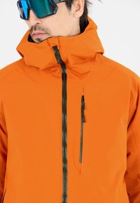 Orange vandtæt jakke med høj krave, frontlynlås og brystlomme. Har en glat tekstur og kontrast sorte lynlåsdetaljer.