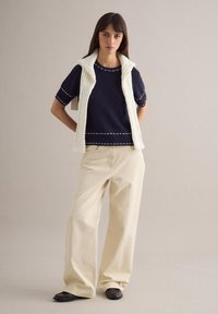 Mujer de pie vistiendo un suéter azul marino de manga corta con costuras blancas, chaleco de punto trenzado color crema sobre los hombros, pantalones anchos color crema y zapatos negros.