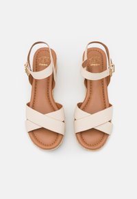 Sandales compensées beiges avec des brides croisées, dessus en cuir lisse, semelle intérieure rembourrée marron et bride de cheville ajustable avec boucle dorée.