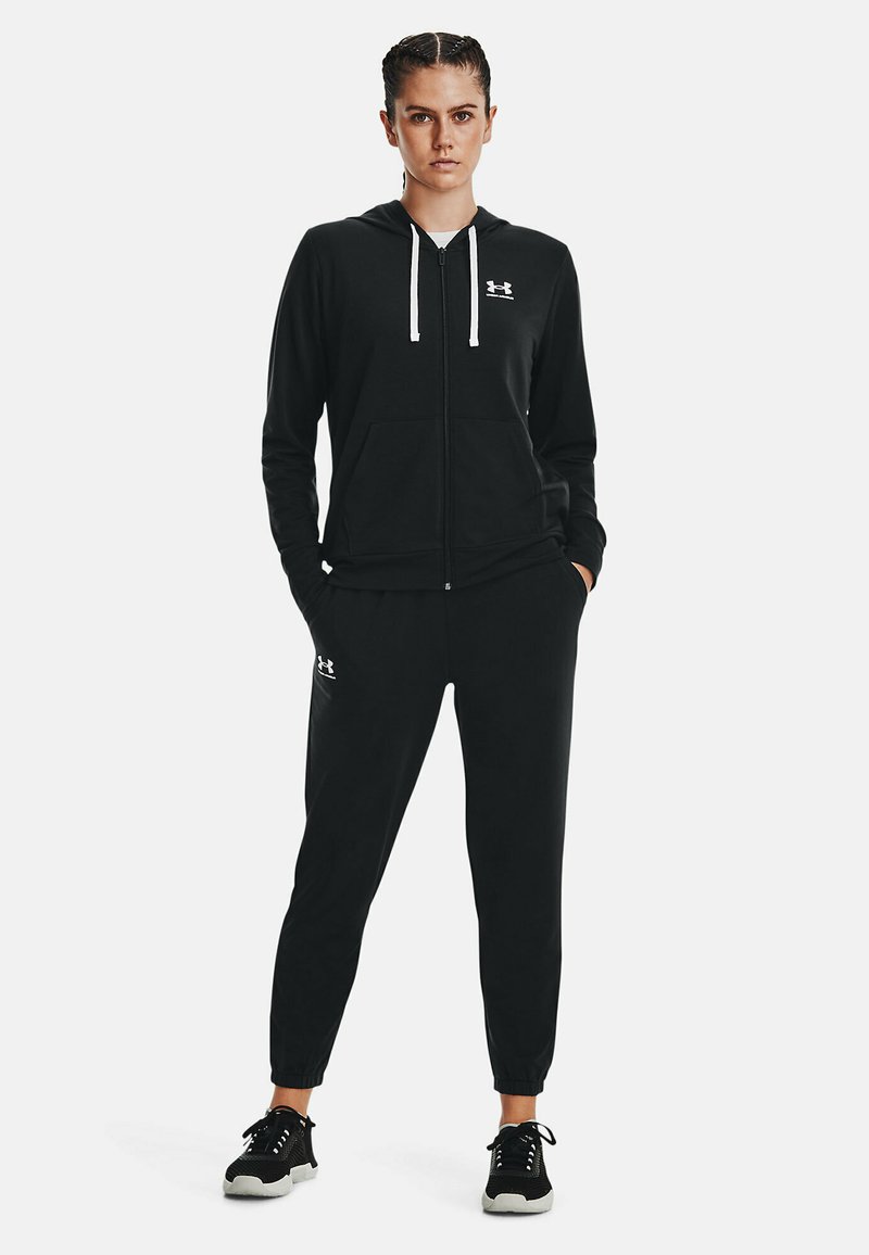 Sudadera con cremallera negra y cordones blancos, combinada con joggers negros. Presenta el logo de Under Armour en ambas piezas. Tejido texturizado, corte holgado.