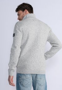 Harmaa neulepusero, jossa on korkea kaulus, teksturoitu ribbikuvio ja ribbikuvioidut hihansuut. Puettuna vaaleansinisten farkkujen päällä.