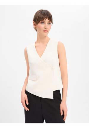 Femme aux cheveux courts bruns portant un gilet blanc asymétrique sans manches et un pantalon noir sur fond blanc.