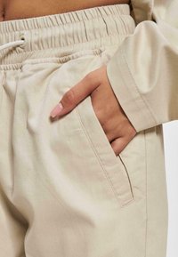 Pantaloni de trening din bumbac bej, cu talie elastică, șnur, buzunar lateral și textură netedă. Vedere în detaliu a unei mâini în buzunar.