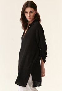 Black button-up shirt in textured fabric, featuring a V-neck, rolled sleeves, and side slits for movement.