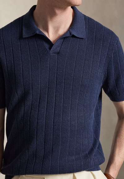 Homme portant un polo en maille bleu marine à manches courtes avec une texture côtelée verticale, associé à un pantalon de couleur claire sur un fond neutre.