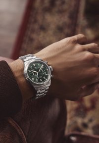 Srebrna kronografska ura iz nerjavečega jekla z temno zelenim obrazom, belimi oznakami ur in brušenim kovinskim zapestjem na zapestju.