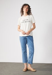Camiseta cremosa con texto negro "LA VIE EST BELLE", combinada con jeans cortos de color azul claro y bailarina de color tostado. Textura suave y corte casual.