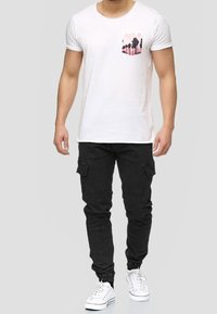 Weißes T-Shirt mit kurzen Ärmeln und pinkem Brusttaschendruck von Palmen, kombiniert mit schwarzen Cargo-Hosen mit Seitentaschen und schmal zulaufenden Knöcheln.