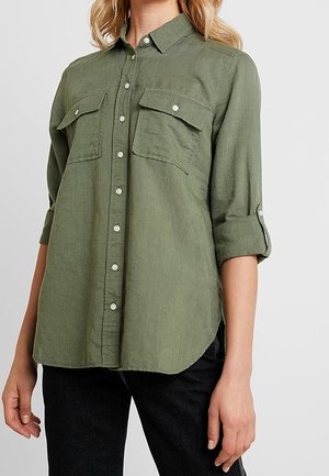 Femme portant une chemise boutonnée vert olive avec deux poches à rabat à l'avant et des manches retroussées, associée à un pantalon noir.