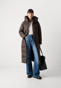 Donkerenbruine pufferjack, knielang, met een hoge kraag en een ritsluiting. Gecombineerd met lichtblauwe high-waisted flared jeans en zwarte schoenen.