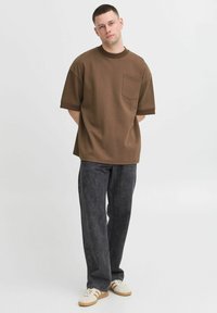 Brun oversized T-skjorte med korte ermer, med lomme, sammen med mørkegrå vide jeans og hvite sneakers med beige detaljer.