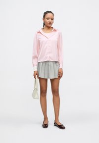 Camisa de cetim cor-de-rosa com botões na frente, minissaia plissada cinzenta e sapatilhas brilhantes cor de vinho, complementada com uma pequena mala branca.