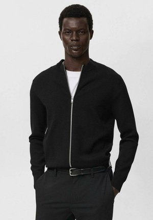Uomo che indossa un maglione nero con zip sopra una camicia bianca e pantaloni neri con una cintura nera, in piedi con le mani nelle tasche.