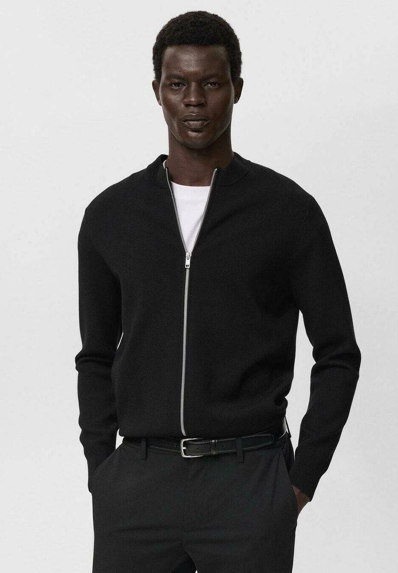 Homme portant un pull zippé noir sur une chemise blanche et un pantalon noir avec une ceinture noire, se tenant les mains dans les poches.