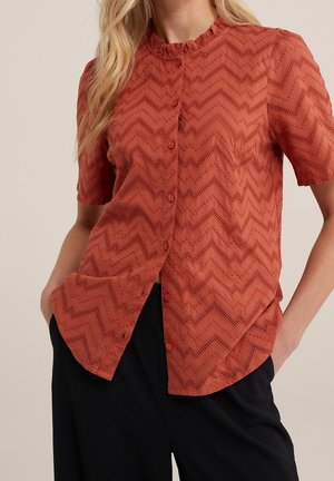 Mujer con blusa de manga corta color óxido con patrones de encaje en zigzag y pantalones negros, manos en los bolsillos, sobre un fondo liso.