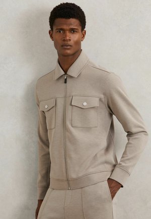 Bēšs rāvējslēdzēja džemperis ar apkakli, divām priekšējām kabatām un ribotām aprocēm. Gluda tekstūra, pieguļošs dizains un minimālistisks stils.