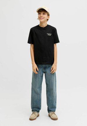 Fant, oblečen v črno majico s tekstom "Originals Selected", modre kavbojke, bež čevlje in bež kapo, stoji in se smeji pred enobarvnim ozadjem.