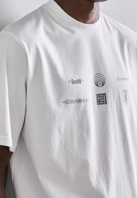 Camiseta de algodón blanca con múltiples logotipos impresos en gris oscuro, incluidos "GmbH" y diseños abstractos, con cuello redondo y mangas cortas.
